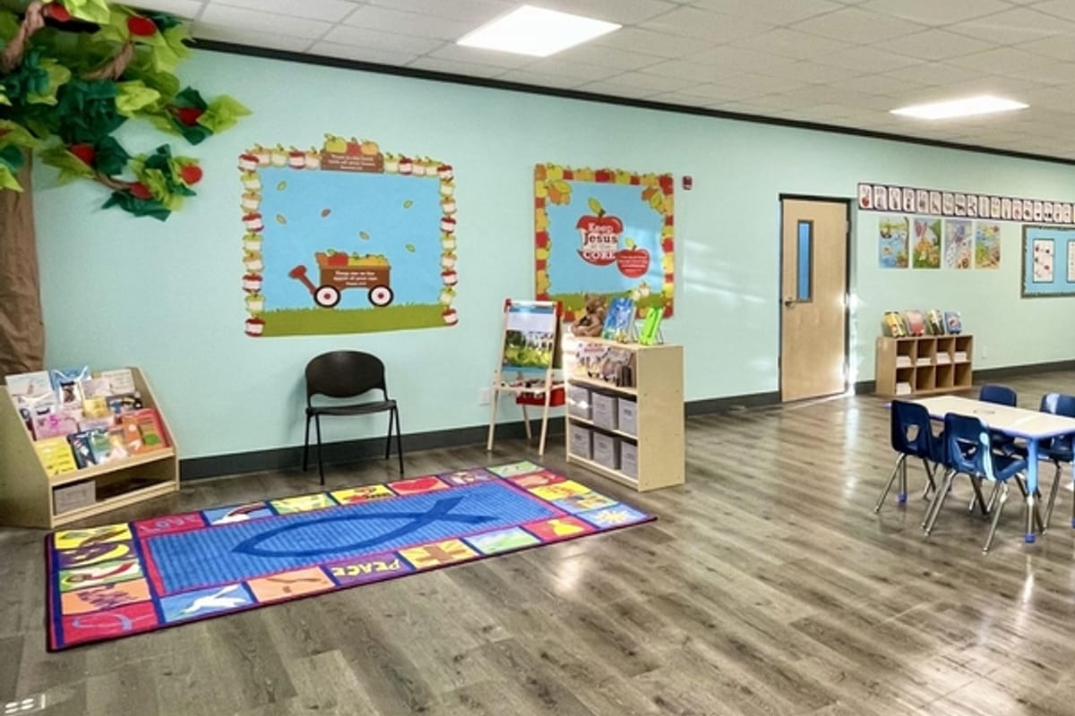 Bright preschool reading corner with a blue-bordered rug, low bookshelves, and wall murals of a wagon and apples; child-sized chair nearby—an inviting learning space.